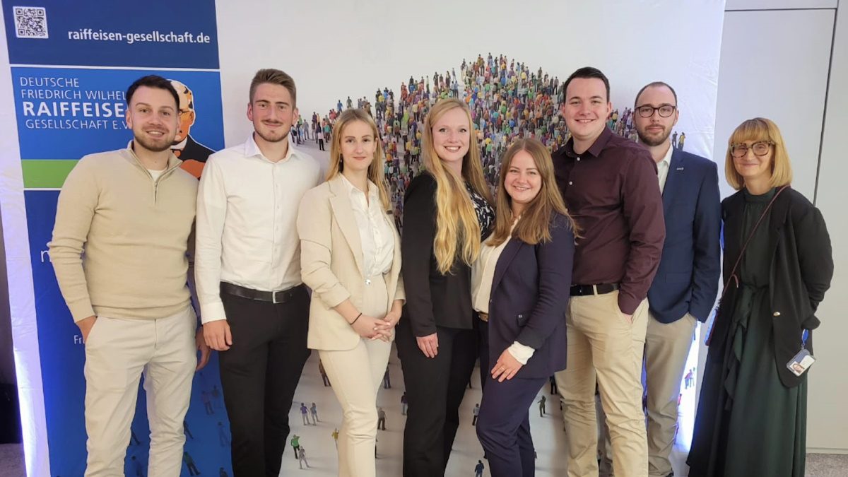 Das Foto zeigt eine Gruppe von Personen, die in formeller Kleidung posieren. Im Hintergrund befindet sich ein Display oder Plakat mit dem Schriftzug "Deutsche Agrar- und Ernährungswirtschaft" und der Marke "Raiffeisen". Außerdem ist ein künstlerisches Bild oder Poster mit vielen kleinen Figuren zu sehen.