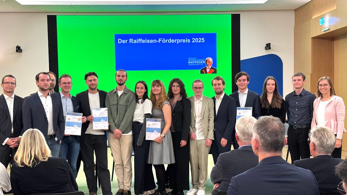 Das Bild zeigt eine Gruppe von Menschen, die zusammenstehen und einige von ihnen halten Auszeichnungen oder Urkunden in den Händen. Im Hintergrund ist eine grüne Wand mit einem hellblauen Banner, worauf "Der Raiffeisen-Förderpreis 2023" steht. Dies scheint eine Preisverleihungsveranstaltung zu sein, bei der die abgebildeten Personen für ihre Leistungen oder Beiträge geehrt werden. Die Teilnehmer scheinen formell gekleidet zu sein, was auf die Bedeutung der Veranstaltung schließen lässt. Im Vordergrund sitzen einige Personen, die offenbar Zuschauer dieses Ereignisses sind.