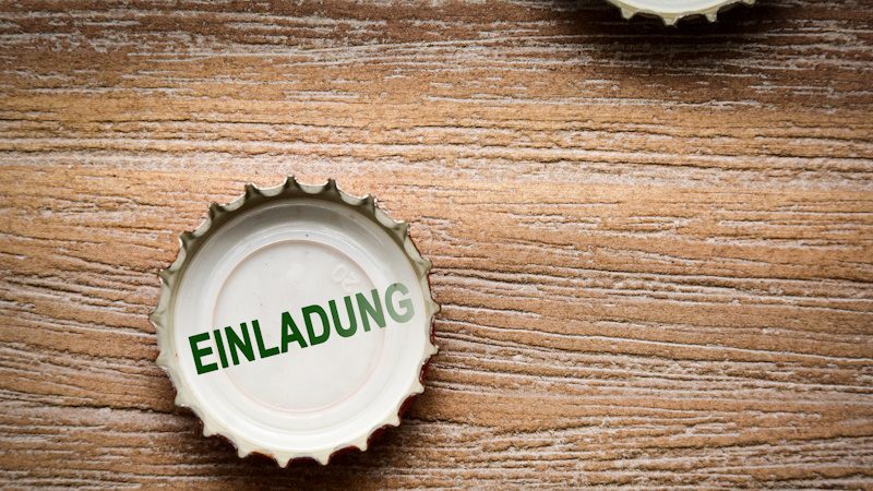 Das Bild zeigt einen Kronkorken, auf dem das Wort "Einladung" steht. Zwei Kronkorken sind auf einer hölzernen Oberfläche platziert. Wahrscheinlich handelt es sich hierbei um eine kreative Darstellung einer Einladung zu einer Veranstaltung, einem sozialen Treffen oder einer Party, bei der Getränke serviert werden. Der Kronkorken symbolisiert die Beziehung zu Getränken, möglicherweise Bier oder Soda, die oft in geselliger Runde verzehrt werden.