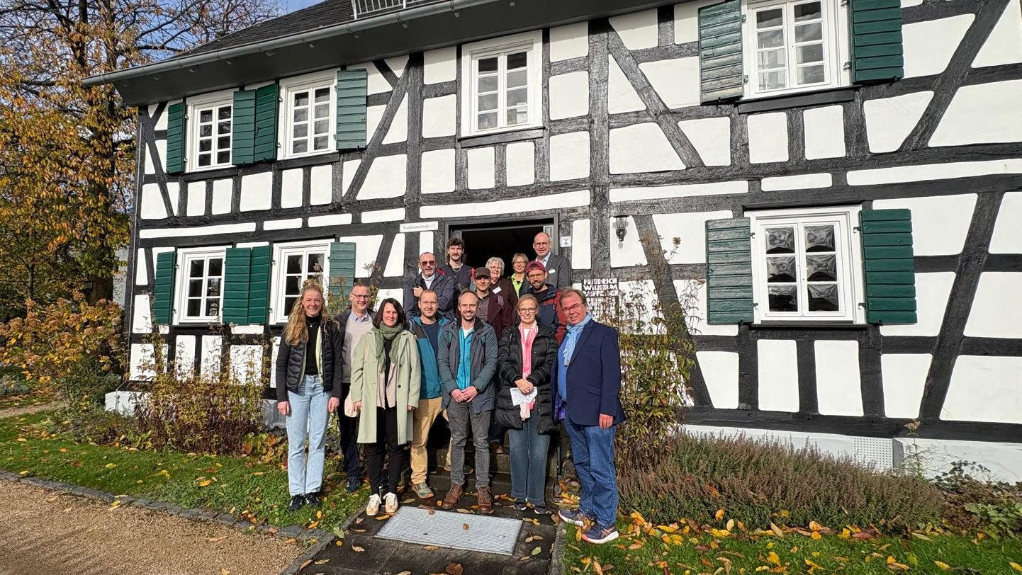 Das Bild zeigt eine Gruppe von Personen, die vor einem Fachwerkhaus stehen. Der Herbst scheint eingetreten zu sein, da die Blätter auf den Boden gefallen sind und einige Bäume am Rand des Bildes ihre Blätter verloren haben. Die Gruppe besteht aus Männern und Frauen verschiedenen Alters, die gemeinsam für das Foto posieren. Das Fachwerkhaus im Hintergrund hat grüne Fensterläden und weiße Wände mit schwarzen Holzbalken, typisch für traditionelle deutsche Architektur. Die Szene vermittelt eine freundliche und gemeinschaftliche Atmosphäre.