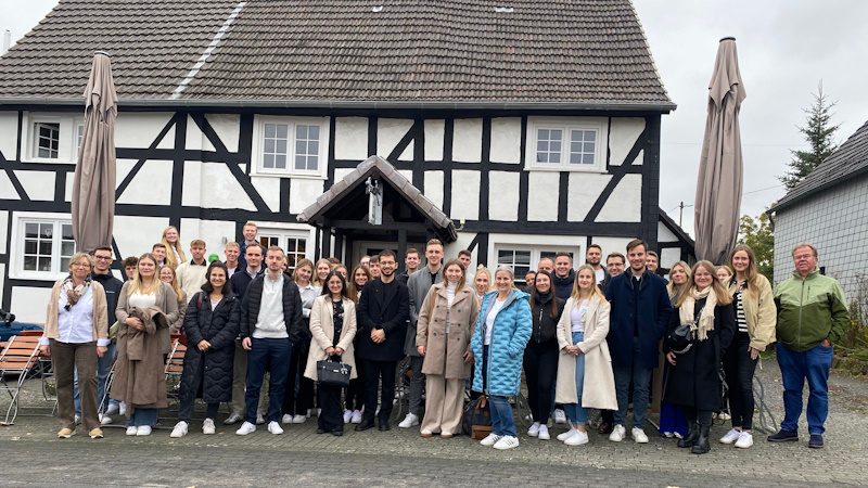Das Bild zeigt eine Gruppe von Menschen, die sich vor einem traditionellen Fachwerkhaus versammelt haben. Fachwerkhäuser sind charakteristisch für bestimmte Regionen in Europa, vor allem in Deutschland, und zeichnen sich durch ihr Holzrahmenwerk, das durch weiße Wände ergänzt wird, aus.

**Beschreibung des Szenarios:**

1. **Gruppenfoto:** Die Personen stehen eng zusammen und posieren für das Foto, was auf eine Art von Gemeinschaft oder gegenseitige Verbundenheit hinweist. Es könnte sich um eine Gruppenveranstaltung oder einen Ausflug handeln.

2. **Kleidung:** Die meisten Menschen tragen Übergangskleidung, wie Mäntel, was auf die Herbst- oder frühe Frühlingszeit schließen lässt.

3. **Atmosphäre:** 
    - **Soziale Zusammenkunft:** Die Atmosphäre wirkt freundlich und entspannt, was typisch für Gruppenbilder bei gesellschaftlichen Ereignissen oder Treffen ist.
    
4. **Umgebung:**
    - **Hintergrund:** Das Haus und die Umgebung könnten ein ländliches Gebiet darstellen, möglicherweise im Rahmen eines touristischen Ausflugs oder von historischen Interessen.
    - **Wetter:** Das Bild ist bei bewölkten Bedingungen aufgenommen worden, was die Jahreszeit und das Klima widerspiegeln könnte.

5. **Persönliche Interaktionen:** Einige Teilnehmer halten Kontakt zu anderen, weisen darauf hin, dass es sich um bekannte oder befreundete Personen handelt.

Dieses Bild könnte Teil eines Berichts über eine Veranstaltung, eine Familie oder eine Gruppenreise sein, bei der Teilnehmer die Schönheit und Architektur traditioneller europäischer Häuser erleben.