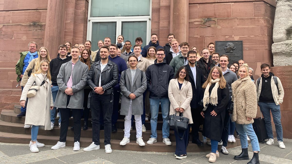 Das Bild zeigt eine Gruppe von Menschen, die offenbar als Team oder Gruppe vor einem Gebäude mit sandsteinfarbenen Wänden stehen. Die Gruppe besteht aus Männern und Frauen unterschiedlichen Alters, die lächeln und sich entspannen, während sie für das Foto posieren. Es könnten Kollegen sein, die bei einem Firmen-Event oder einem Teambuilding-Ausflug zusammenkommen. Die Kleidung variiert von leger bis künstlerisch-schick, mit einigen Personen in Jacken oder Mänteln, was auf kühleres Wetter hindeuten könnte. Das Gebäude hinter ihnen scheint historisch oder öffentlicher Natur zu sein, was möglicherweise darauf hindeutet, dass die Gruppe sich zum Zwecke der Weiterbildung, eines Meetings oder aus einem feierlichen Anlass dort versammelt hat.