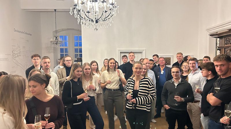 Das Bild zeigt eine Gruppe junger Menschen, die sich in einem elegant eingerichteten Raum versammelt haben. Einige von ihnen halten Getränke in der Hand und es scheint eine gesellige Veranstaltung oder ein Empfang zu sein, wo die Teilnehmer miteinander plaudern und sich amüsieren. Im Hintergrund ist ein Kronleuchter und große Fenster zu sehen, die dem Raum eine stilvolle Atmosphäre verleihen. Auf der linken Seite befindet sich eine Wand mit einer Zeichnung oder Aufschrift, die sich jedoch nicht klar erkennen lässt.