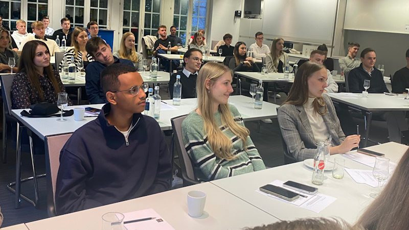 Ein Gruppenmeeting oder Seminar in einem Konferenzraum mit mehreren jungen Erwachsenen, die aufmerksam der Präsentation oder dem Gespräch im Raum zuhören. Die Tische sind mit Notizblöcken, Schreibutensilien und Getränken ausgestattet.