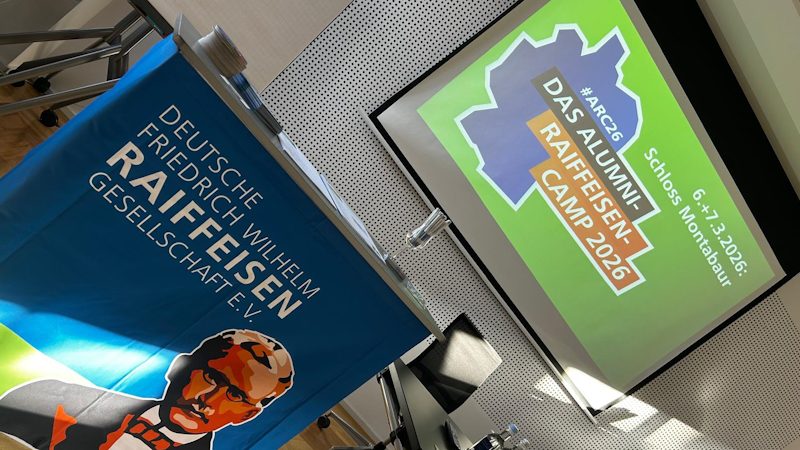 Das Bild zeigt eine Veranstaltung oder Präsentation der "Deutschen Friedrich Wilhelm Raiffeisen Gesellschaft e.V.". Es gibt ein Banner mit dem Bild von Friedrich Wilhelm Raiffeisen und einen Projektor, der das "Raiffeisen Camp 2026" ankündigt. Die Veranstaltung findet im Schloss Montabaur statt, vom 6. bis 7. Februar 2026.