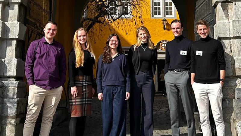 Das Bild zeigt eine Gruppe von sechs jungen Erwachsenen, die zusammen vor einem historischen Gebäude mit einem steinernen Bogen stehen. Sie sind lächelnd und tragen unterschiedliche formelle Kleidung. Hinter ihnen ist eine gelbe Wand und ein Baum zu sehen.