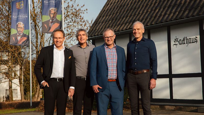 Das Bild zeigt mehrere Personen, die vor einem Gebäude mit der Aufschrift "Backhaus" stehen. Im Hintergrund sind Flaggen mit Raiffeisen zu sehen.